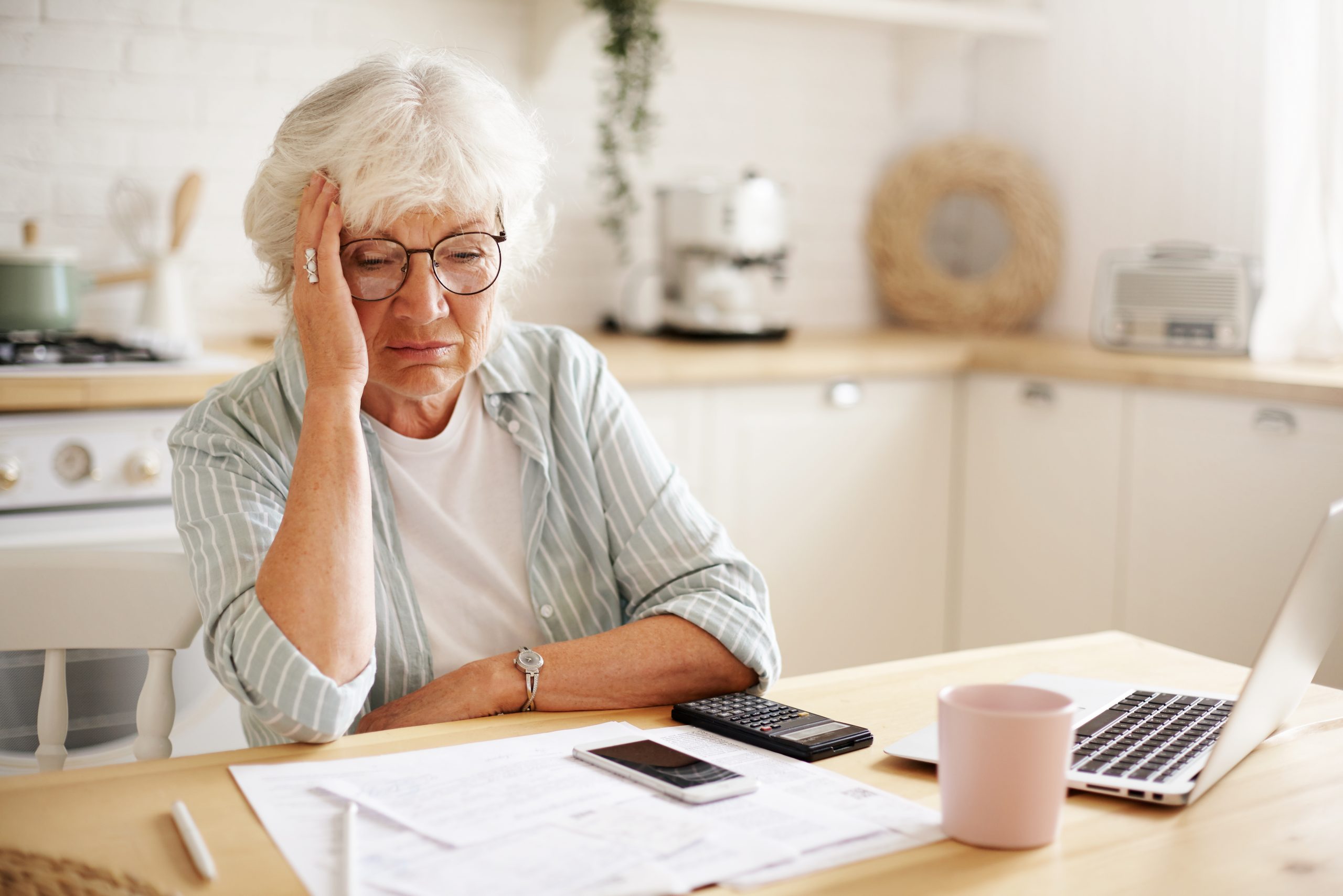 Sad frustrated senior woman pensioner having depressed look, holding hand on her face, calculating family budget, sitting at kitchen counter with laptop, papers, coffee, calculator and cell phone Medicare Frustrations